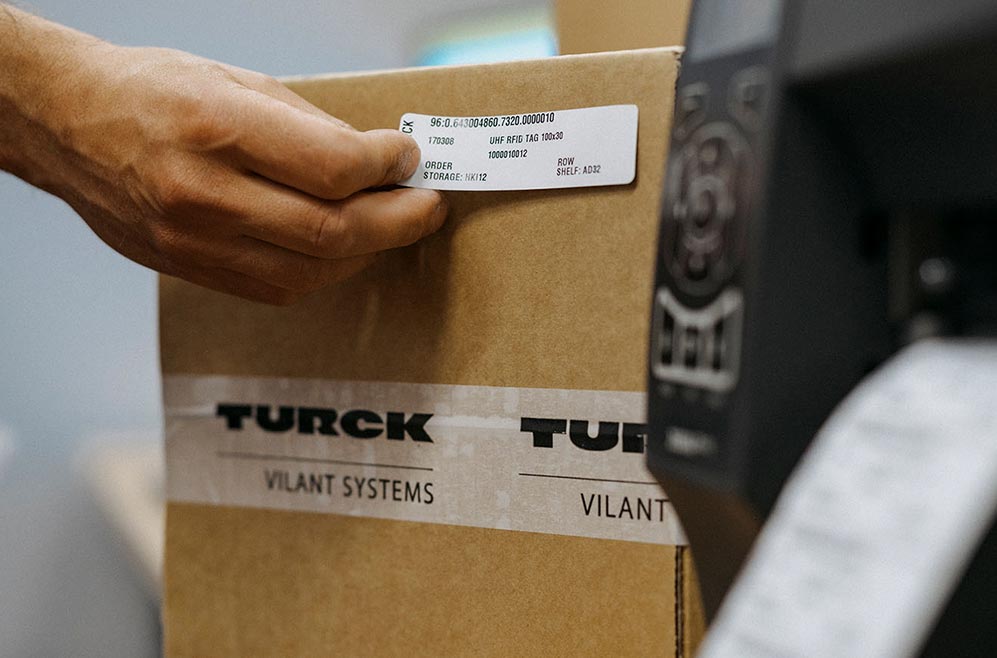 A box and a printer plus a person's hand attaching a label on the side of the box.