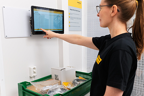 A person using a tablet PC on the wall with a crate of items on a table depicting Scan Room of RFID self service