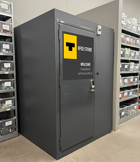 A picture of a wholesale facility where shelves are fully stocked. Focus of the picture is a checkout room or cage, whose entrance is visible. The checkout room serves different self-service solutions with RFID